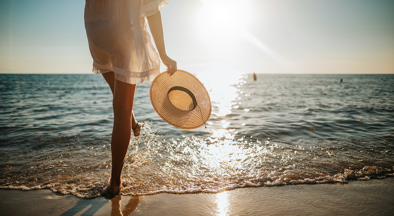 woman on a beach near the shoreline with a hat in her hand looking out at the water